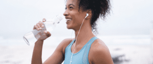 A woman with headphones drinking water while standing on the beach.