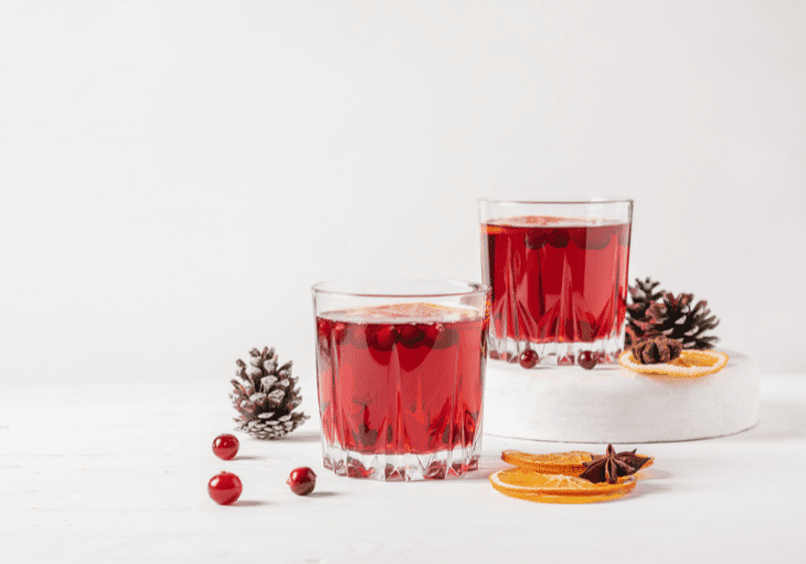 Two glasses of cranberry juice next to a pine cone and orange.