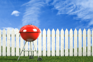 A red grill sitting in the grass next to a white fence.